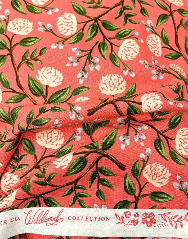 May include: Coral pink fabric with a white floral pattern. The fabric features a repeating design of white flowers with green leaves and stems. The fabric has a white label with the text "Wildwood Collection" printed on it.
