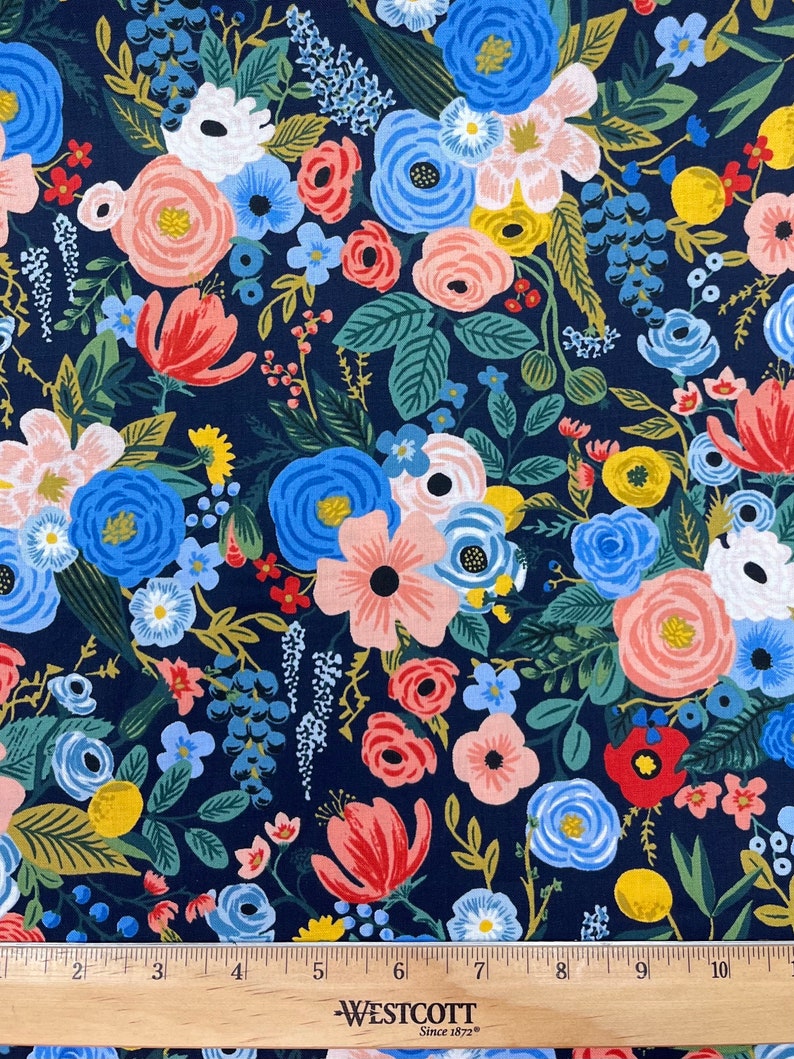 May include: A navy blue fabric with a floral pattern featuring pink, red, blue, and yellow flowers. The fabric is printed with a variety of flowers and leaves, creating a busy and colorful design. The fabric is shown with a ruler, indicating that it is sold by the yard.