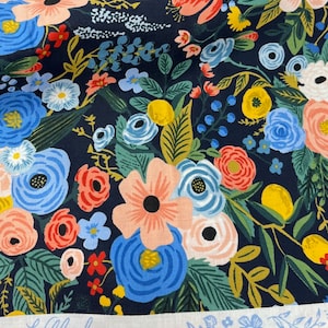 May include: A navy blue fabric with a floral pattern featuring pink, red, blue, yellow, and green flowers. The fabric has a white label with the text "CO. Wildwood COLLECTION".