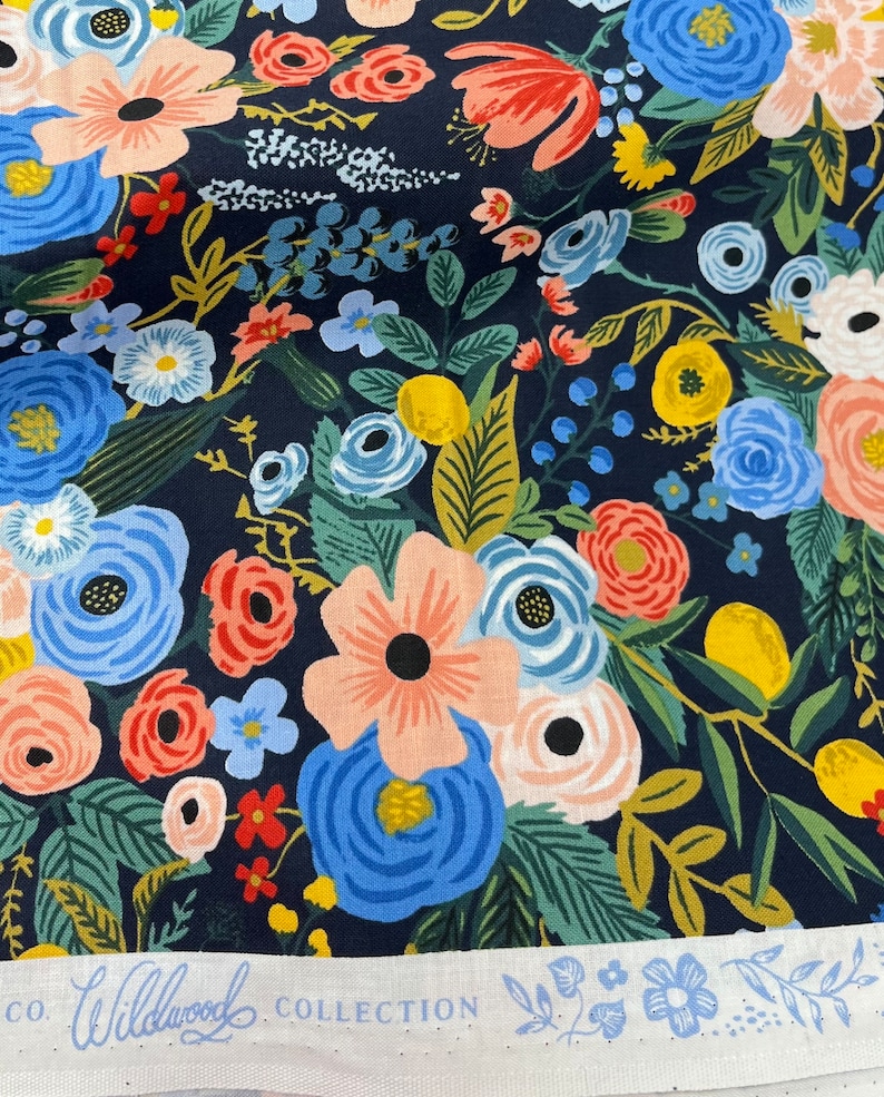 May include: A navy blue fabric with a floral pattern featuring pink, red, blue, yellow, and green flowers. The fabric has a white label with the text "CO. Wildwood COLLECTION".