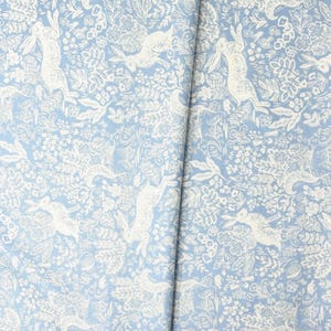 May include: Light blue fabric with a repeating pattern of white rabbits, flowers, and foliage. The design creates a whimsical, nature-inspired aesthetic. The fabric is likely cotton or linen, suitable for various sewing projects.
