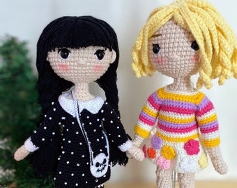 May include: Two crocheted dolls, one with black hair and a black and white polka dot dress, the other with blonde hair and a striped shirt and a white skirt with colorful polka dots. Both dolls are wearing pink shoes. A crocheted hand is lying on a wooden surface.