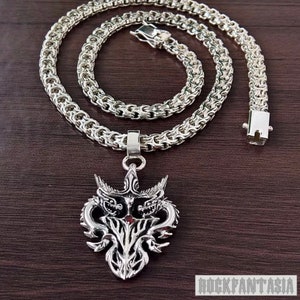 Puede incluir: Un collar de cadena de plata con un colgante de plata que presenta un diseño detallado de dos dragones con una piedra preciosa roja en el centro. El colgante está unido a la cadena con un cierre de plata. El collar está colocado sobre una superficie de madera.