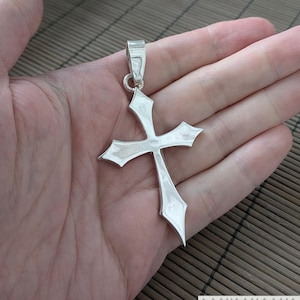 Può includere: Un ciondolo a forma di croce argentata con bordi appuntiti, tenuto in una mano aperta. Il ciondolo ha un anello nella parte superiore per una catena. La croce è di colore argento brillante.