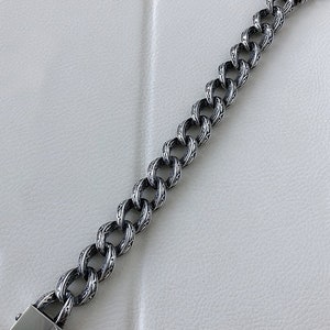 K&ouml;nnte beinhalten: Ein silbernes Kettenarmband mit einem detaillierten, komplizierten Design. Das Armband hat einen Verschluss.