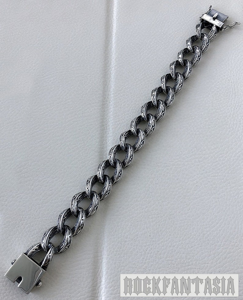 K&ouml;nnte beinhalten: Ein silbernes Kettenarmband mit einem detaillierten, komplizierten Design. Das Armband hat einen Verschluss.