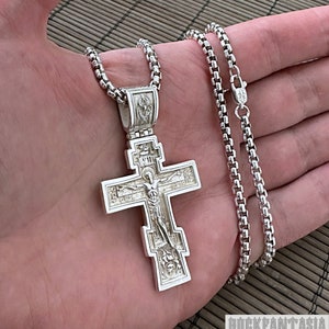 Puede incluir: Un colgante de cruz ortodoxa de plata con una detallada representación de Jesucristo en una cadena de collar tipo caja. La cruz presenta grabados intrincados.