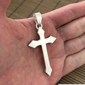 Può includere: Un ciondolo d'argento a forma di croce. La croce presenta un design dettagliato con estremità appuntite e un anello per una catena. Il ciondolo è mostrato su uno sfondo neutro.