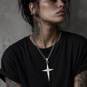 May include: A silver pendant necklace with a four-pointed star design. The pendant is suspended from a chain. The person in the image is wearing a black t-shirt.