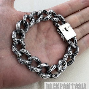 K&ouml;nnte beinhalten: Ein silbernes Kettenarmband mit einem detaillierten, komplizierten Design. Das Armband hat einen Verschluss.