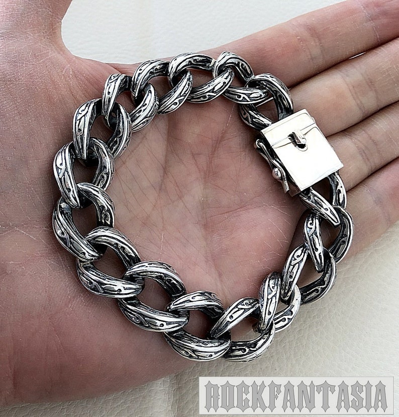 K&ouml;nnte beinhalten: Ein silbernes Kettenarmband mit einem detaillierten, komplizierten Design. Das Armband hat einen Verschluss.