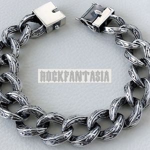 K&ouml;nnte beinhalten: Ein silbernes Kettenarmband mit einem detaillierten, gravierten Muster. Das Armband hat einen Verschluss.
