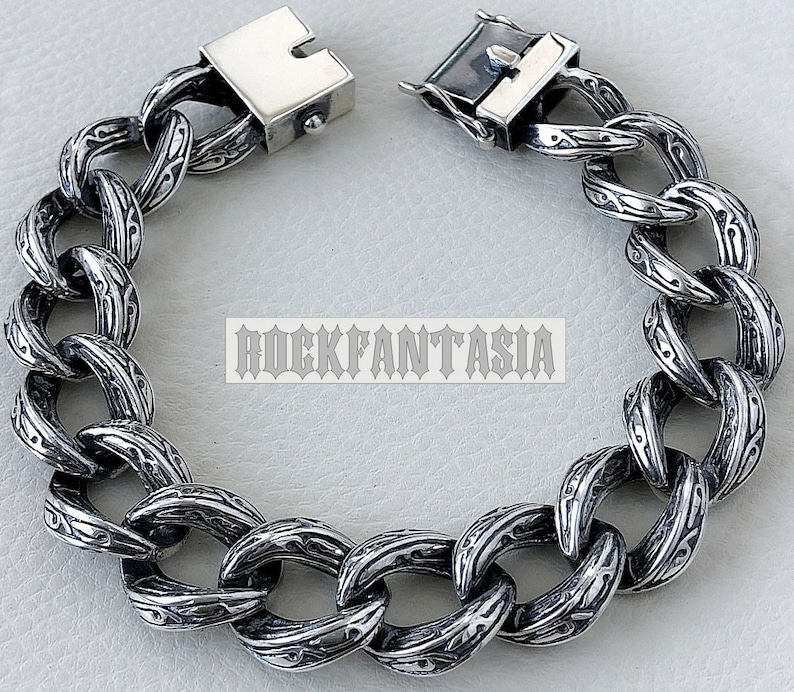 K&ouml;nnte beinhalten: Ein silbernes Kettenarmband mit einem detaillierten, gravierten Muster. Das Armband hat einen Verschluss.