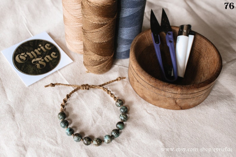 May include: A turquoise and brown beaded bracelet with a brown cord. The bracelet is tied with a knot and has a simple design. The bracelet is on a white fabric surface with other craft supplies.