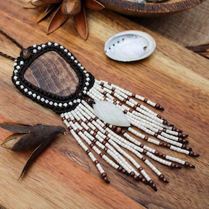 May include: A handmade necklace featuring a rectangular wooden pendant, edged with black and white beads. The pendant has a white, leaf-shaped stone and beaded fringe. The necklace is displayed on a wooden surface.