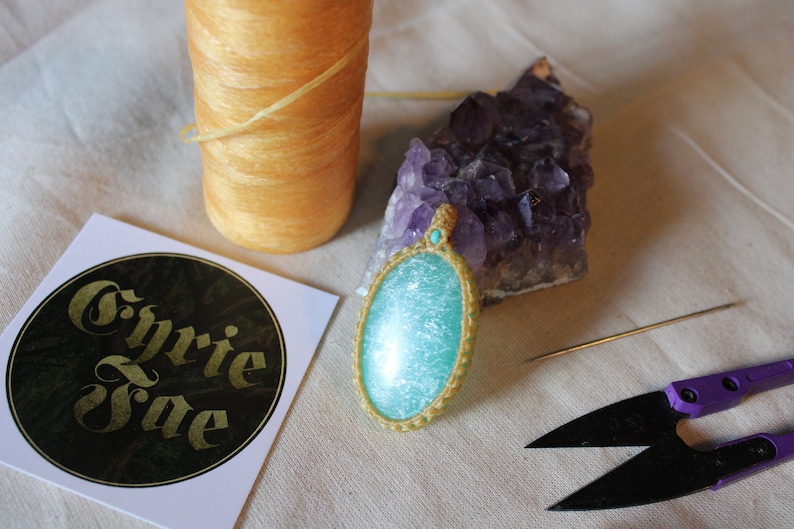 May include: A turquoise and gold pendant with a sparkly finish, a spool of yellow thread, a needle, a pair of purple handled scissors, and a sticker with the text "Cyrie Jae" on a white background.