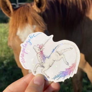May include: A sticker featuring a unicorn illustration with a rainbow mane and tail. The word "unapologetic" is written in cursive. The sticker has a holographic finish and is set against a blurred background.