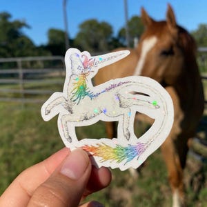May include: A white unicorn sticker with a rainbow mane and tail. The sticker has a holographic, glittery effect. The unicorn is held in front of a brown horse in a field.