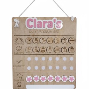 May include: Wooden daily responsibility chart for "Clara", with pink and white details. Includes sections for morning and evening tasks, completed tasks, daily stars, and rewards. Features a hanging rope.