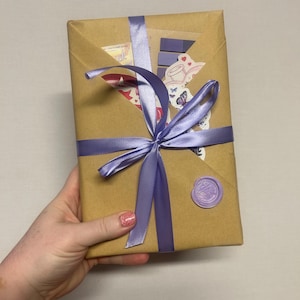 May include: A wrapped gift in brown paper, tied with a lavender satin ribbon and bow. The package is sealed with a lavender wax seal. Decorative stickers are visible on the front of the package.