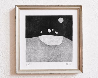 May include: Framed black and white art print. The print shows a minimalist landscape with a dark sky, a moon, and a light-coloured ground. The print is in a white matted frame with a silver-coloured wooden frame. The print is labelled "PK '24" and "1/60".