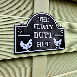 May include: A black and white sign reading "THE FLUFFY BUTT HUT" with chicken illustrations. The sign is mounted on a light green, ridged surface and secured with two silver screws.