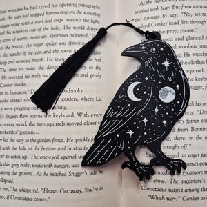 Puede incluir: Marcapáginas negro con forma de cuervo con un diseño celestial de estrellas, una luna creciente y un planeta. El marcapáginas tiene una borla negra y está colocado en un libro abierto. El número de página 176 es visible.