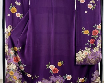 May include: A vibrant purple kimono with floral patterns. The garment features a detailed design of colorful flowers, including red, yellow, and white blossoms, along the edges and sleeves. The kimono is displayed on a black clothing rack.