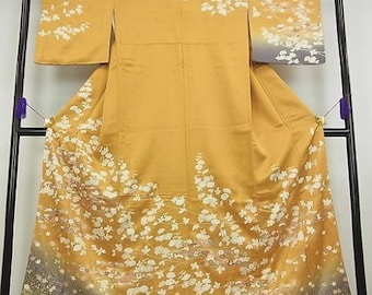 Könnte beinhalten: Ein traditioneller japanischer Kimono mit goldfarbenem Hintergrund und einem weißen Blumenmuster. Der Kimono wird auf einem schwarzen Ständer ausgestellt.