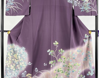May include: A purple silk kimono with a floral pattern. The kimono features a design of white flowers, green leaves, and gold accents. The flowers are arranged in a circular pattern on the lower part of the kimono, while the leaves are arranged in a more linear pattern on the upper part of the kimono.