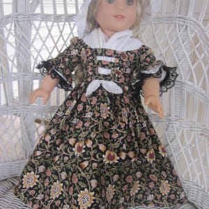 May include: A doll wearing a black floral print dress with a white collar and cuffs. The dress features a pattern of yellow, pink, and green flowers. The doll also wears a white bonnet with lace trim. The doll is seated on a white wicker chair.