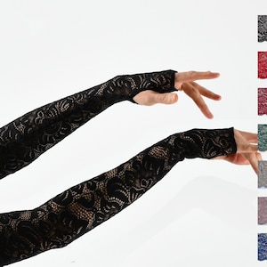 Puede incluir: Un par de guantes sin dedos de encaje negro. Los guantes se muestran en los brazos de una modelo, con un brazo extendido y el otro doblado en el codo. Los guantes están hechos de un delicado tejido de encaje con un patrón floral.