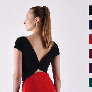 Op de afbeelding: Een zwart, kort, mouwloos topje met een gedraaid detail op de rug. Het topje is getailleerd en heeft een diepe V-hals. Het model draagt het topje met een rode rok.