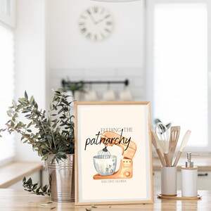 May include: A framed print with a watercolor illustration of a kitchen mixer with the text "Feeding the patriarchy" and "Soli Deo Gloria" in black lettering.