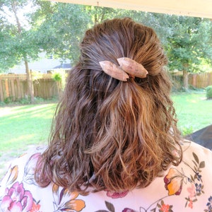 May include: A woman with brown, wavy hair is wearing a floral print blouse. Her hair is pulled back with a pair of brown, wooden hair clips.