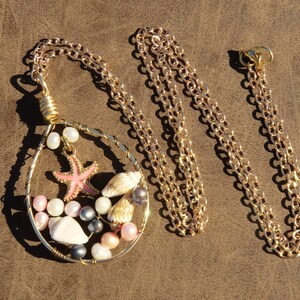 May include: A gold-colored necklace with a pendant featuring a pink starfish, seashells, and pearls. The pendant is wire-wrapped and hangs from a gold chain. The necklace is displayed on a brown surface.