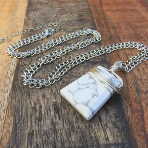May include: A white howlite stone pendant wrapped in silver wire, hanging from a silver chain necklace. The necklace is on a wooden surface.
