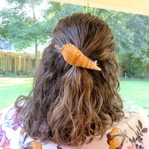 May include: A woman with long brown hair is wearing a seashell hair clip. The clip is a light brown color and has a natural, organic shape. The woman is wearing a floral patterned shirt.