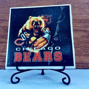 May include: A ceramic tile with a black background featuring a cartoon image of a bear wearing a football jersey with the text "CHICAGO BEARS". The tile is on a black metal stand.