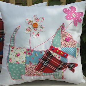 Può includere: Un cuscino bianco con un design a patchwork di un cane con un collare rosso a quadri e un fiore rosa. Il cane è fatto di diversi tessuti, tra cui stampe floreali, quadri e vichy.