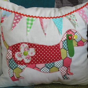 May include: A white pillow with a red and white polka dot dachshund sewn on. The dachshund is made of various patterned fabrics, including a red and white polka dot fabric, a green and yellow honeycomb fabric, and a floral fabric. The pillow has a red scalloped trim with a row of colorful fabric flags sewn on top.
