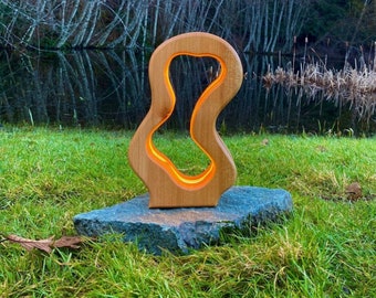 Könnte beinhalten: Eine Holzlampe mit einer einzigartigen, organischen Form. Die Lampe ist beleuchtet und strahlt ein warmes Licht aus. Sie steht auf einem Stein in einem grasbewachsenen Bereich.