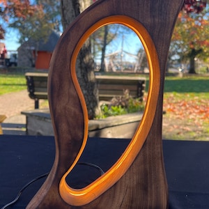 May include: A dark brown wooden lamp with a unique, abstract design. The lamp has a warm, glowing light that shines through the cutout in the wood.