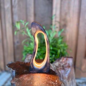 May include: A wooden lamp with a distinctive, natural form. The dark brown wood has a central, illuminated aperture that gives off a warm light. The lamp is placed on a natural wood base, creating a rustic and artistic aesthetic. The lamp's height is approximately 20 cm.