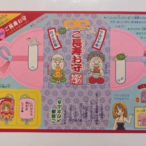 May include: A Japanese paper craft with illustrations of elderly figures, turtles, and cranes. The text includes "Longevity Amulet" and "Eyeglass Cleaner Case". The design features pink, blue, and yellow elements.