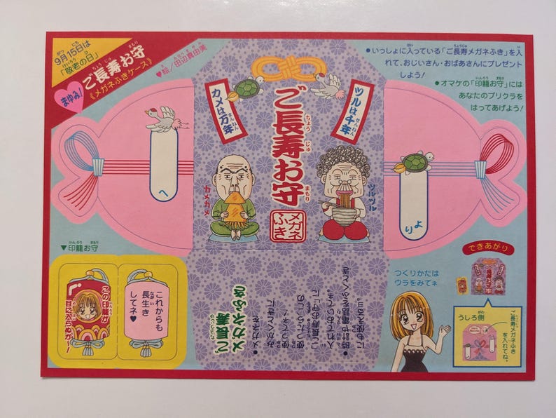 May include: A Japanese paper craft with illustrations of elderly figures, turtles, and cranes. The text includes "Longevity Amulet" and "Eyeglass Cleaner Case". The design features pink, blue, and yellow elements.