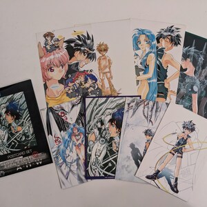 Può includere: Un set di cartoline in stile anime con vari personaggi dai capelli blu, rosa e neri. Il set include una cartellina nera con la scritta POSTCARD SET e ZION, contenente diverse schede rettangolari che mostrano illustrazioni dettagliate di figure in pose dinamiche.