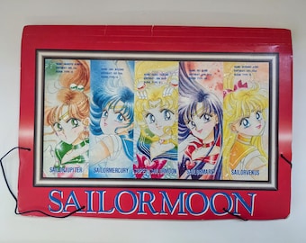 Op de afbeelding: Een rode map met een zilveren frame met een kleurrijke illustratie van vijf anime-personages uit de Sailor Moon-serie. De personages zijn Sailor Jupiter, Sailor Mercury, Super Sailor Moon, Sailor Mars en Sailor Venus. De tekst "Sailor Moon" is in blauwe letters op de voorkant van de map gedrukt.