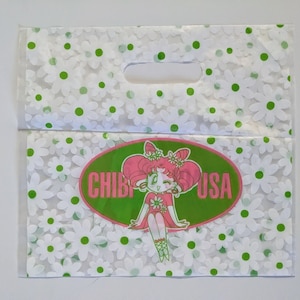 May include: A white plastic shopping bag with a green and white daisy pattern. The bag has a handle and a logo that says "CHIBI USA" with a cartoon girl in a pink dress.
