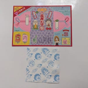 May include: A Japanese souvenir set. The set includes a pink and purple illustrated card with cartoon characters and text, and a white cloth printed with blue cartoon faces.